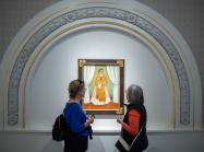 Two museum visitors are viewing a painting. The painting features a woman with a medium skin-tone wearing a dress and scarf and holding a piece of paper. It is framed by an arc with golden and floral ornamentation.