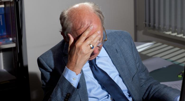 Florian Van Roeke photograph of a bald man in a blue suit at his desk with his head in his hand